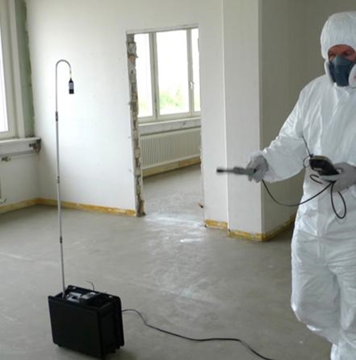 Person in protective gear uses a device to measure air quality in an empty room.